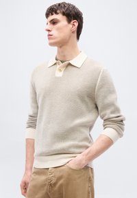 Νεαρός άνδρας που φοράει μπεζ πουλόβερ με υφή πλεκτού, γιακά και κουμπιά, συνδυασμένο με ανοιχτό καφέ παντελόνι, ποζάροντας με το ένα χέρι στην τσέπη.