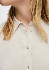 Linen button-up shirt in light beige, featuring a classic collar, five buttons, and a textured fabric with a subtle grid pattern.