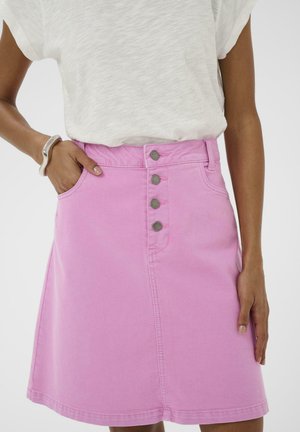 Persoană purtând o fustă roz deschis cu nasturi în față, cu mâna în buzunar, și un top alb cu mâneci scurte, pe un fundal simplu.