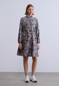 Φόρεμα με λεοπάρ μοτίβο, εμπρόσθιο φερμουάρ, μακριά μανίκια, ζωνάκι στη μέση με κορδόνι και τσέπες. Φορεμένο με λευκά αθλητικά παπούτσια πάνω σε ουδέτερο φόντο.