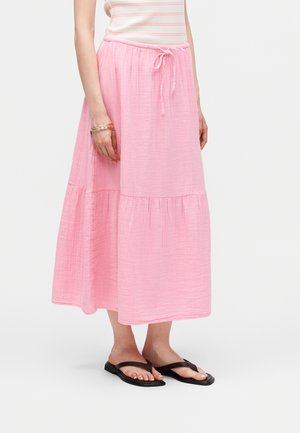 Žena nosící růžskou vrstvenou sukni až na kotníky s nastavitelným pasem, černé sandály na rovné noze a zlaté náramky na levém zápěstí.