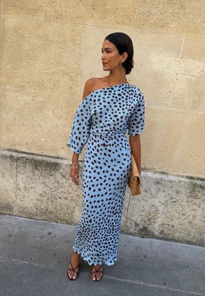 Femme debout à l'extérieur portant une robe bleue à une épaule avec des taches noires, tenant une pochette marron et portant des sandales marron à lanières.