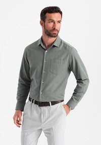 Chemise verte à boutons avec une poche poitrine, manches longues et boutons bleus, associée à un pantalon gris clair et une ceinture marron.