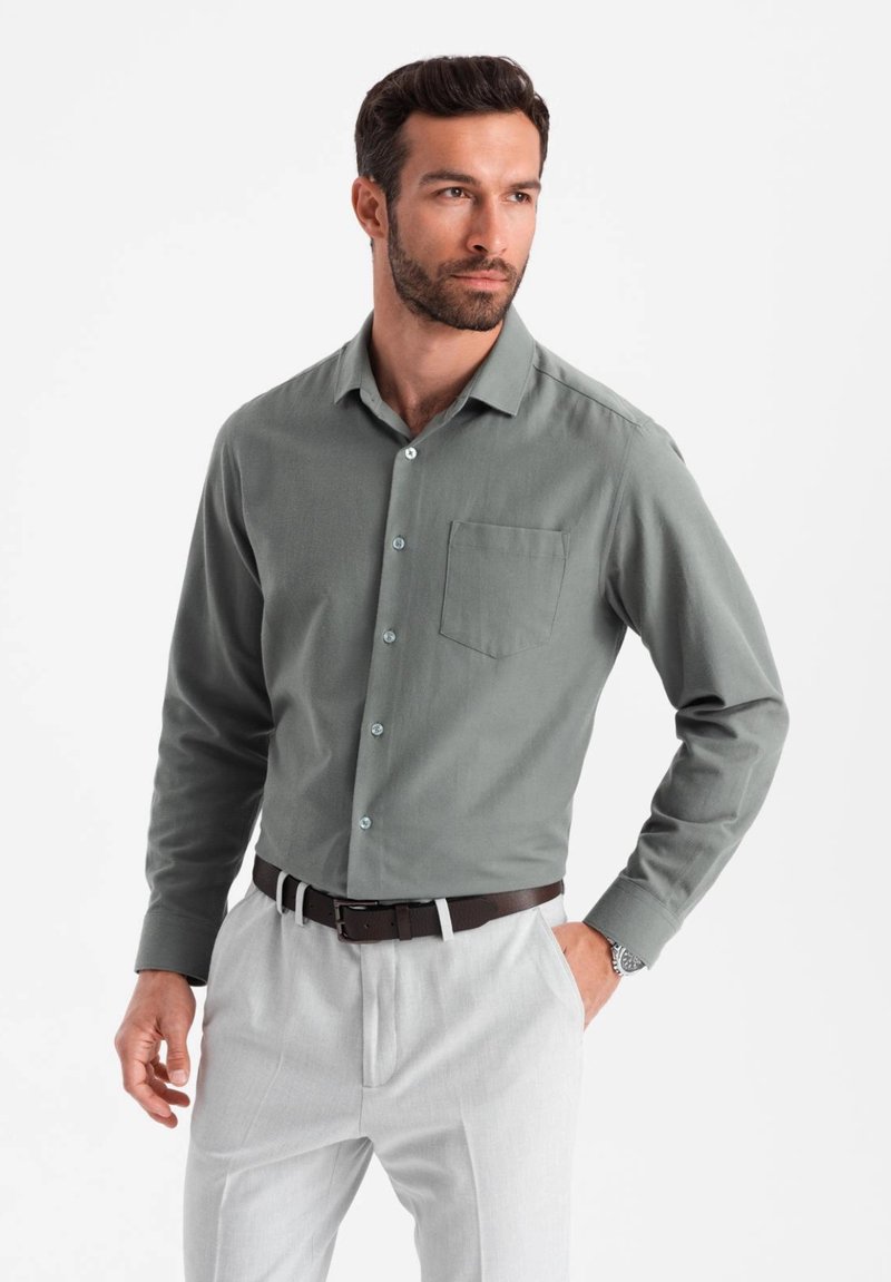 Chemise verte à boutons avec une poche poitrine, manches longues et boutons bleus, associée à un pantalon gris clair et une ceinture marron.