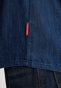 Jeans in denim blu con taglio dritto e un'etichetta di tessuto rossa con "JOOP! JEANS" cucita sul lato vicino all'orlo.