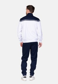 Witte ritsjack met marineblauwe accenten, hoge kraag en lange mouwen, gecombineerd met marineblauwe broek en witte sneakers. Effen textuur, geen patronen.