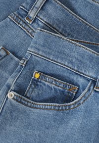 Gros plan d'un jean en denim bleu montrant la poche avant avec des rivets métalliques et des détails de couture orange-brun.