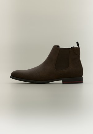 Bottine Chelsea en daim marron avec panneaux latéraux élastiques noirs et semelle noire avec une bande rouge, montrée de profil sur un fond neutre.