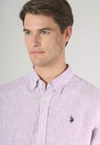 Un uomo con capelli corti e acconciati indossa una camicia a righe rosa e bianca con un piccolo logo navy sul petto, guardando leggermente fuori dalla camera.