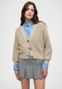 Beige, strikket cardigan med V-hals, tre knapper og ribbestrikket kant, sammen med en blå, stripete skjorte med knapper og en grå, foldet skjørt.