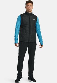 Gilet isolé noir avec un col montant et une fermeture éclair à l'avant, superposé à une chemise à manches longues bleue. Un pantalon noir et des baskets complètent la tenue.