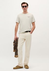 Hellbeiges T-Shirt mit Grafik, cremefarbene Slim-Fit-Hose, brauner gestreifter Pullover über dem Arm und braune Lederloafers. Schwarze Sonnenbrille.