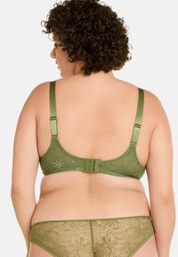 Soutien-gorge en dentelle verte avec bretelles réglables, présentant des motifs floraux et une fermeture à agrafe. Thong assorti en dentelle verte transparente visible.