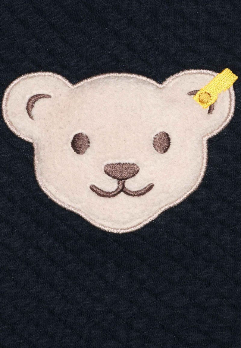 Weißes Filz-Bären-Gesicht mit braunen Akzenten auf einem dunklen, gesteppten Hintergrund. Besitzt runde Ohren, gestickte Augen und ein gelbes Stoffetikett.