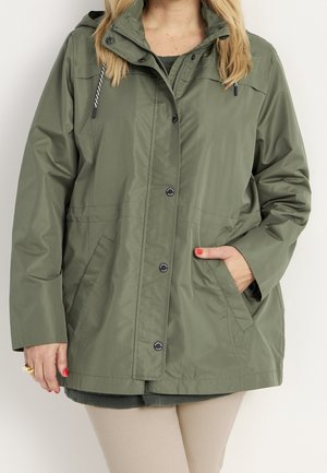 Frau trägt eine olivgrüne Kapuzenjacke mit Druckknöpfen, Seitentaschen und Kordelzug, kombiniert mit beigen Hosen.