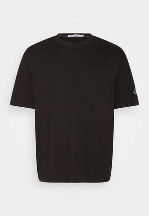 T-shirt en coton noir avec col rond et manches courtes. Détail notable : petit patch logo sur la manche gauche.