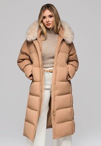 Cappotto imbottito beige con cappuccio foderato in pelliccia, silhouette lunga, texture trapuntata e zip frontale. Indossato su un maglione lavorato a maglia beige e pantaloni bianchi.