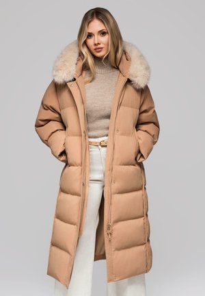 Abrigo acolchado beige con capucha forrada de piel, silueta larga, textura acolchada y cremallera frontal. Lleva puesto sobre un suéter de punto beige y pantalones blancos.