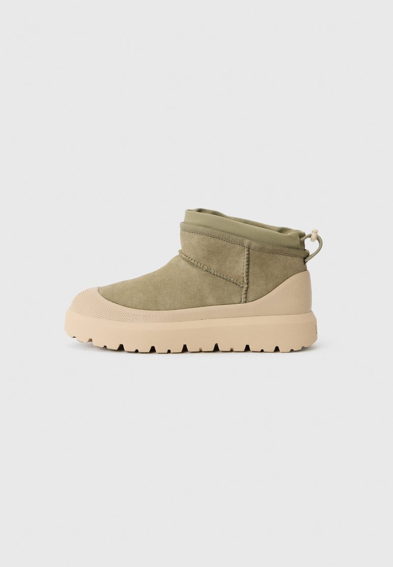 Kaki mockaskinnankelboot med en beige gummisula och ett omgivet elastiskt topp. Funktioner en dragflik på baksidan för enkel påtagning och texturerade detaljer.