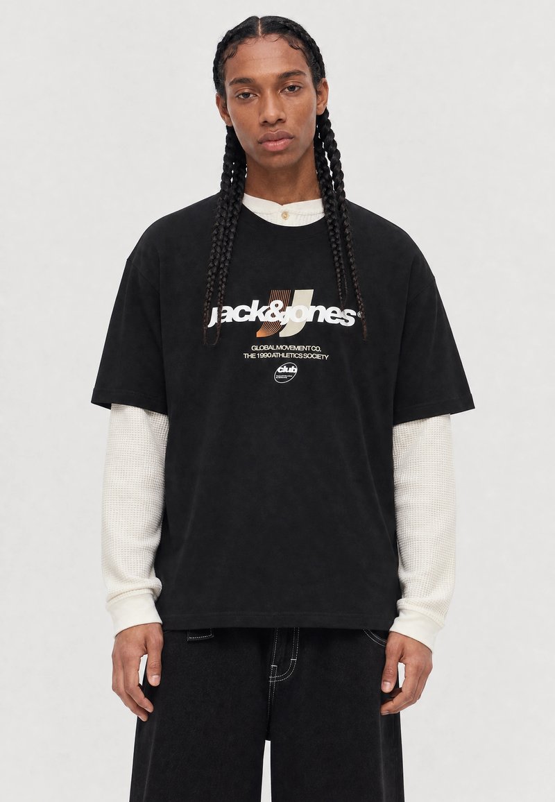 Άτομο με μακριά πλεξούδα φορά στρωτό μαύρο T-shirt Jack & Jones και λευκό πουκάμισο με μακριά μανίκια σε απλό φόντο.
