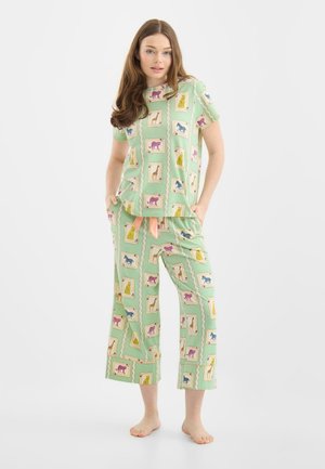 Kobieta stojąca boso, ubrana w jasnocytrynowy komplet piżamy z kolorowymi zwierzęcymi nadrukami, w tym żyrafami, końmi i dużymi kotami, z rękami w kieszeniach.