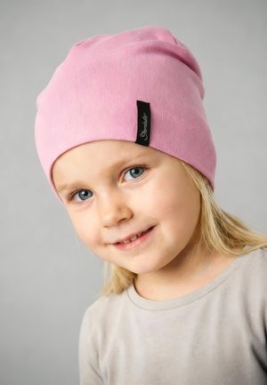 Young child with blue eyes and blonde hair wears a pink beanie with a black tag, smiling slightly, against a plain gray background.