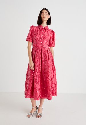 Kvinne iført en rosa blomstret midikjole med puffede ermer, frontknapper, belte i livet og sølvfargede høyhælte sandaler som står på hvit bakgrunn.