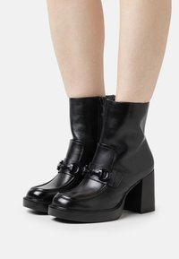 Bottines en cuir noir avec un talon carré, présentant une texture lisse, une fermeture éclair et une sangle décorative avec détail en métal.