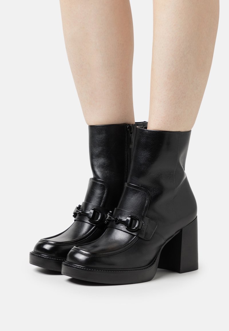 Bottines en cuir noir avec un talon carré, présentant une texture lisse, une fermeture éclair et une sangle décorative avec détail en métal.