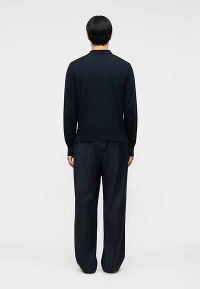 Άνδρας με κοντά μαύρα μαλλιά, που φοράει μαύρο μακρυμάνικο πουκάμισο και φαρδιά, σκούρα παντελόνια με λεπτές ρίγες, στέκεται γυρισμένος με την πλάτη σε απλό φόντο.