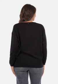 Donna con lunghi capelli castani, che indossa un maglione nero largo e jeans blu sbiaditi, in piedi e girata di spalle contro uno sfondo bianco.