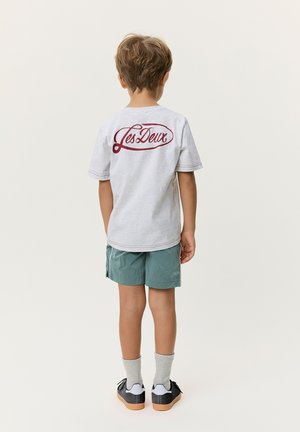Barn med lysebrunt hår står med ryggen til, iført hvid T-shirt med rød "Les Deux" tekst, turkis shorts, grå sokker og sorte sneakers.