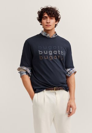 Junger Mann mit lockigem Haar trägt ein schwarzes T-Shirt mit Bugatti-Logo über einem hochgekrempelten karierten Hemd, weiße Hosen und einen geflochtenen braunen Gürtel.