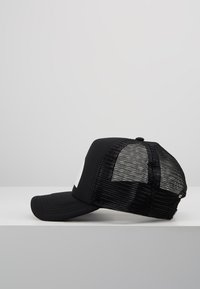 Casquette trucker noire en mesh avec panneau avant rigide et visière courbée, posée sur une surface blanche devant un fond gris.