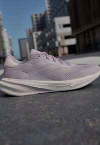Zapatillas de deporte de color púrpura claro con una parte superior de malla texturizada, diseño de tres franjas y una suela blanca curvada, que exhiben un estilo moderno para correr.