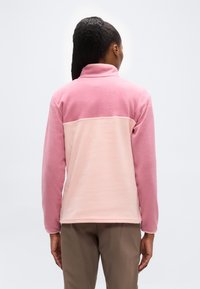 Άτομο με σκούρα πλεξούδα που φοράει διχρω μο ροζ φλις μπουφάν και καφέ παντελόνι, εμφανίζεται από πίσω με φόντο ένα απλό φόντο.