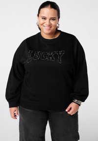 Sweatshirt noir avec "LUCKY" en sequins, doté d'un col rond et de manches bouffantes. Associé à un pantalon gris foncé et des accents argentés.