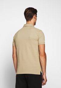 Beige polotröja i en bomullsmix, med krage, korta ärmar och sidofickor för rörelse. Bakvy visas.