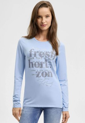 Frau mit langen braunen Haaren, die ein hellblaues Langarmshirt mit motivierendem Text und eine blaue Jeans trägt, vor weißem Hintergrund stehend.