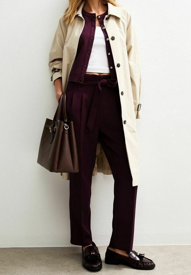 Femme portant un trench-coat couleur crème, un pantalon violet foncé avec ceinture nouée, un cardigan assorti, un crop top blanc, des mocassins noirs, et tenant un sac à main marron foncé.
