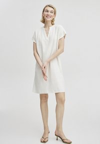 Weißes Leinenkleid mit kurzen Ärmeln, V-Ausschnitt und lockerer Passform. Kombiniert mit grünen, hochhackigen Sandalen, hat es ein einfaches, minimalistisches Design.