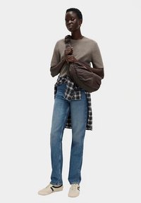 Brązowa puchowa torba, beżowy sweter, koszula w kratę związana w pasie, niebieskie dżinsy i kremowe trampki. Model stoi na jednolitym tle.