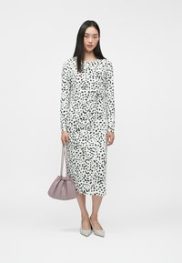 Robe midi blanche à manches longues avec un motif à pois noirs ; design froncé. Associée à un sac violet pâle et des escarpins gris clair.