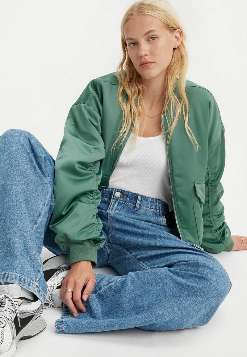 Grüne Satin-Bomberjacke mit Reißverschlussfront, gerippten Bündchen und seitlichen Taschen. Kombiniert mit hellblauen, hochgeschnittenen Weitbeinhosen und Sneakers.