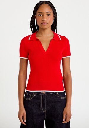 Frau mit geflochtenem Haar, die ein rotes, kurzärmliges Poloshirt mit weißem Rand und schwarze, hoch taillierte Jeans vor weißem Hintergrund trägt.