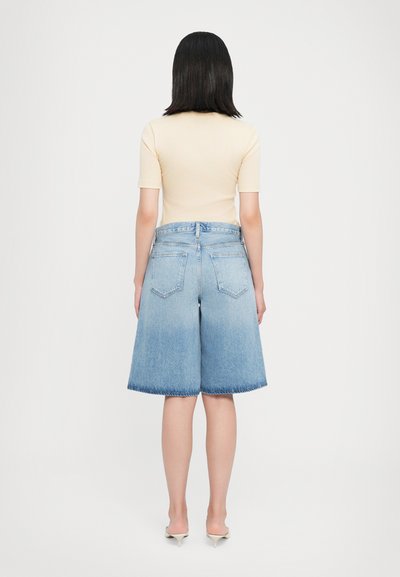 Shorts en denim bleu clair avec un ourlet effiloché, taille mi-haute et deux poches arrière, associés à un haut beige ajusté et des talons de couleur claire.