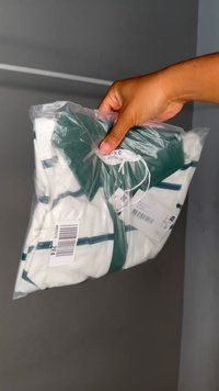 Käsi hoiab selge plastikakotti, mis sisaldab volditud valget särki rohelise krae ja roheliste ning siniste triipudega, müügitagide nähtavusega.