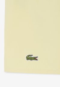 Lichtgeel stof met een klein, groen, geborduurd krokodillenlogo vlakbij de onderrand.