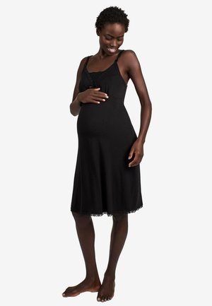 Schwangere Frau in einem schwarzen ärmellosen Kleid, die zärtlich ihren Bauch hält, lächelnd, barfuß vor einem schlichten weißen Hintergrund stehend.