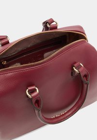 Bordeauxfarbene Ledertasche mit goldfarbener Reißverschluss, doppelten Griffen und strukturierten Oberflächen. Das Innere verfügt über eine mit Logo-Muster gefütterte Auskleidung und eine Reißverschlusstasche.